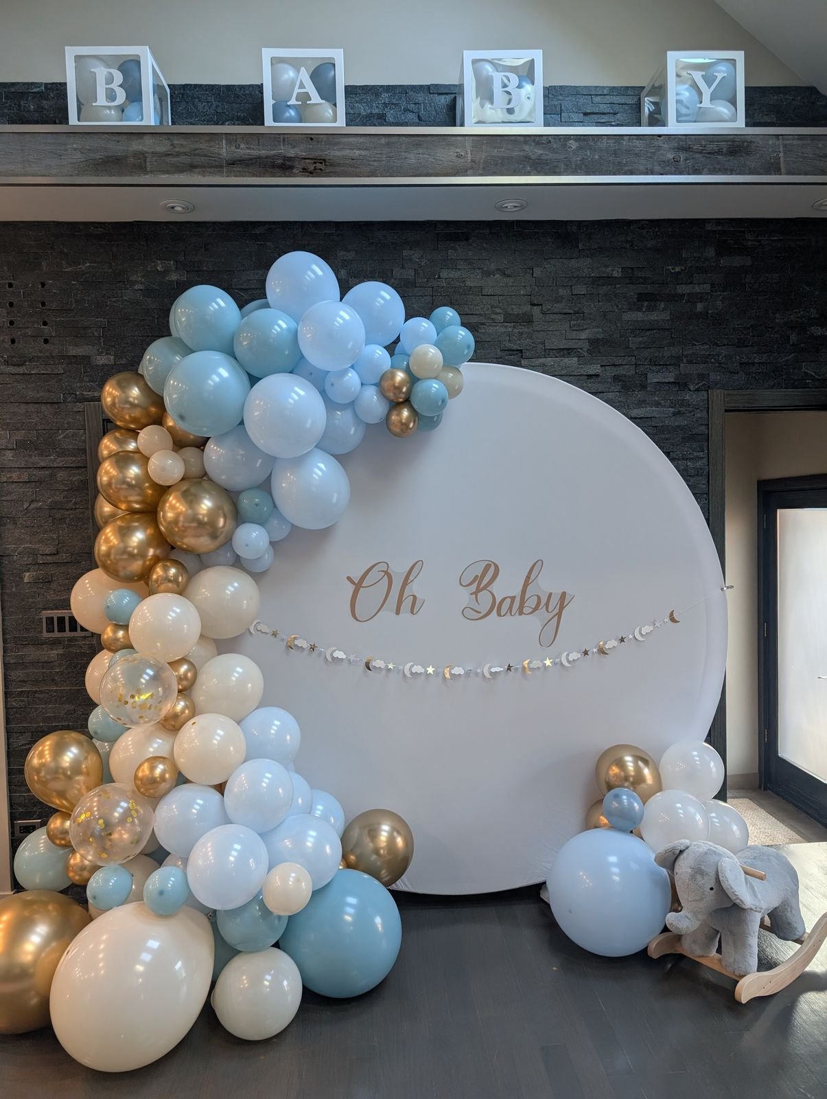 Blue, white and gold balloon garland around a round 'Oh Baby' backdrop